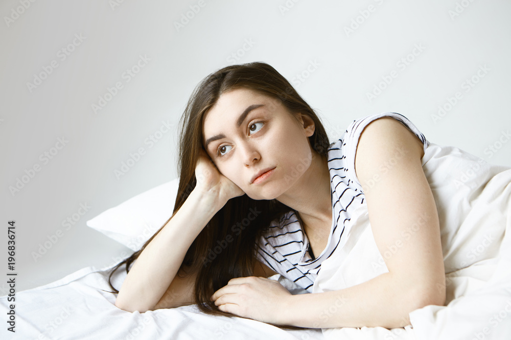 People and lifestyle concept. Beautiful young brunette European woman in striped pajamas feeling lonely and bored to death while lying in bed, looking sideways with thoughtful facial expression People and lifestyle concept. Beautiful young brunette European woman in striped pajamas feeling lonely and bored to death while lying in bed, looking sideways with thoughtful facial expression