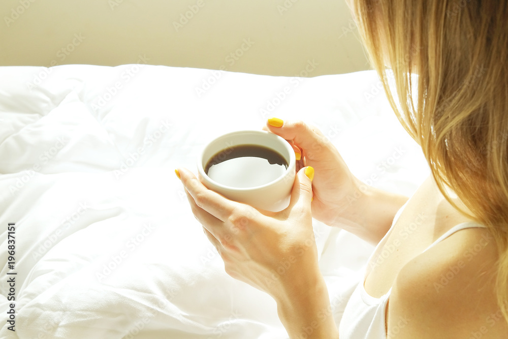 New day new life concept. Young fit woman in big bed w/ white linens sheets holding morning coffee cup. Attractive female wearing sexy tight nightie shirt, bask in sun. Hands close up background