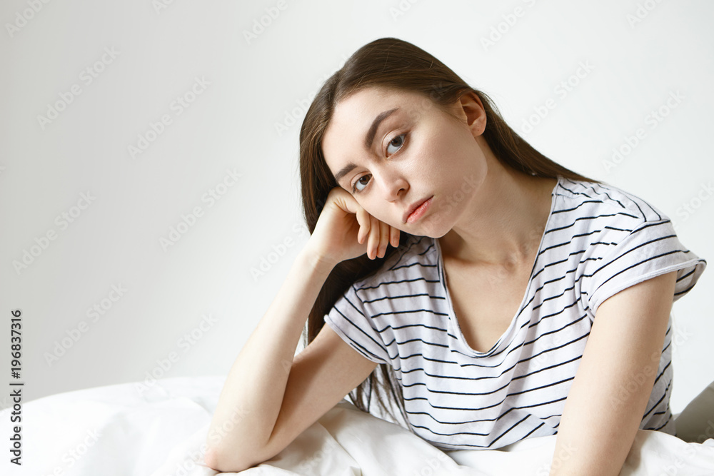 Pretty girl in striped pajamas having bored unhappy expression, looking at camera with hand under her cheek while sitting on bed at white wall with copy space for your text or advertising content Pretty girl in striped pajamas having bored unhappy expression, looking at camera with hand under her cheek while sitting on bed at white wall with copy space for your text or advertising content