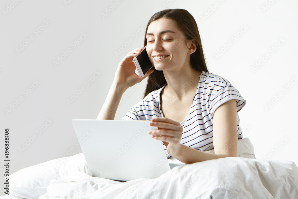 Attractive positive young female in casual wear sitting in bedroom with portable computer and having phone conversation with her boyfriend, smiling happily and closing eyes, glad to hear his voice Attractive positive young female in casual wear sitting in bedroom with portable computer and having phone conversation with her boyfriend, smiling happily and closing eyes, glad to hear his voice