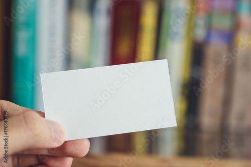 A hand holding empty white business card on wooden table and blur bookshelf in background © Farknot Architect
