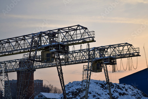 Industrial area. Outdoor crane trestle. Electric overhead traveling crane above the open warehouse and loading area at dawn. A smoking pipe in the background