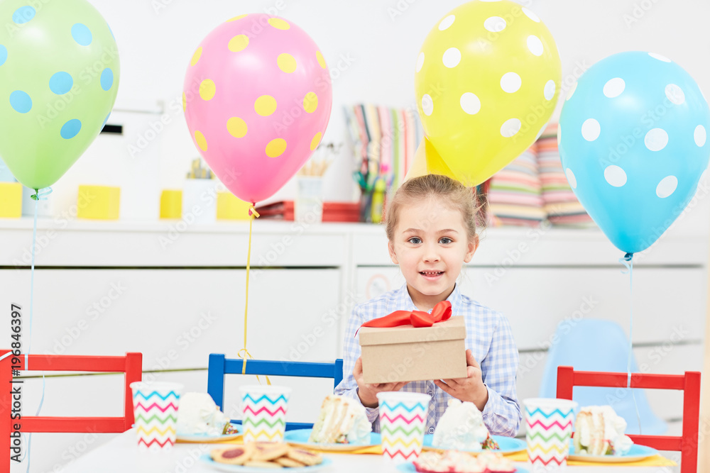 Fototapeta premium Pretty little girl showing box with red ribbon containing her birthday gift while sitting by festive table