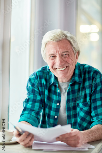 Cheerful aged man with papers looking at camera while spending time in cafe