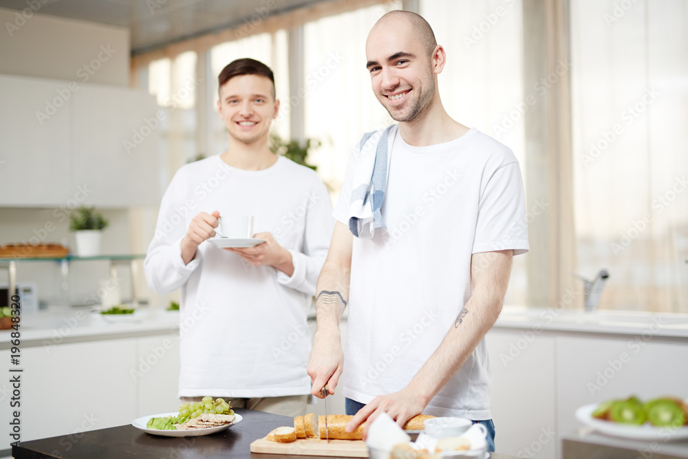 Happy young man cutting baguette for breakfast while standing by table in the kitchen Happy young man cutting baguette for breakfast while standing by table in the kitchen