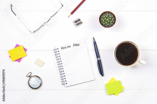 Notepad with the text: action plan. White table with clock, cactus, paper for notes, coffee mug, pen, glasses.