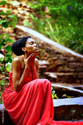 African American girl in a red dress  with dreadlocks sitting on a background of green plants on the rocks in the Park. She closed her eyes  she turned my face to the sun. Retro processing