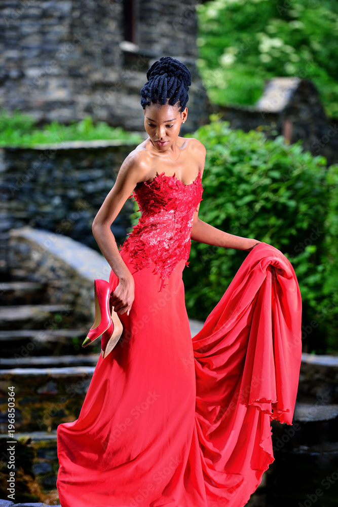 African American girl in a red dress with red shoes in his hand posing in the Park on a background of green trees. Stands on rocks and looks down. Vertical orientation