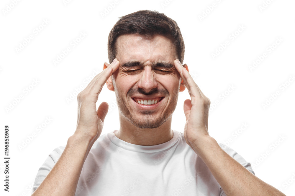 Portrait of young man isolated on white background suffering from severe headache, pressing fingers to temples, closing eyes to relieve pain with helpless face expression Portrait of young man isolated on white background suffering from severe headache, pressing fingers to temples, closing eyes to relieve pain with helpless face expression
