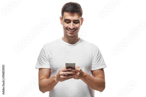 Young man in blank tshirt standing isolated on white background looking attentively at screen of cellphone, browsing web pages and smiling nicely while chatting with friends