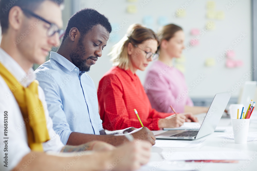 Co-working managers making notes while sitting in row in office or boardroom