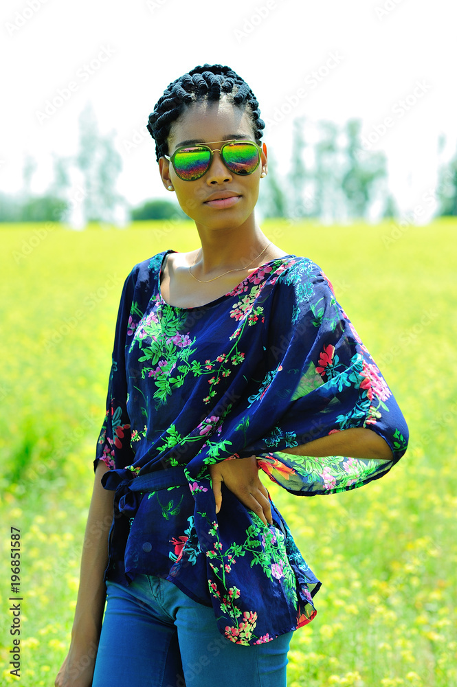 African American girl with dreadlocks, wearing sunglasses, jeans and a tunic, standing in a field of yellow flowers on a Sunny day and smiles