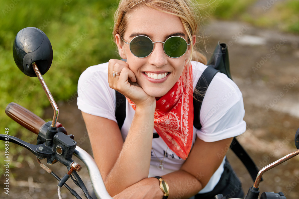 Obraz premium Close up shot of glad stylish motorcyclist being pleased to have outdoor journey on motorbike, carries bag, poses on fast bike alone. Blonde happy woman glad to be involved in extreme driving