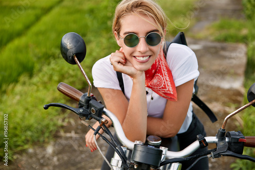 People, transportation and lifestyle concept. Happy young blonde woman dressed casually, being satisfied after fast ride on motorbike, wears trendy sunglasses, dreams about something pleasant