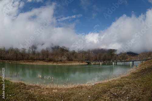 Forest lake with bridge during the sunny day with winter trees and blue cloudy sky. Beautiful natural mountain lake with forest in the background and stormy clouds on the sky.
