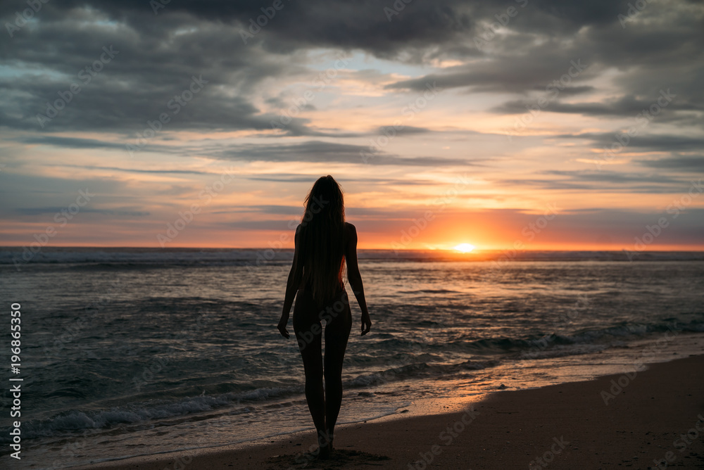 Back view silhouette of sexual woman. Beautiful young girl with long hair walking on the beach, posing at sunset. Concept of travel, relax, yoga