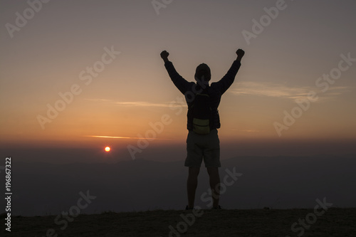 Man with backpack putting his hands up and standing on cliff at sunset time
