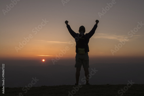 Man with backpack putting his hands up  and standing on cliff at sunset time