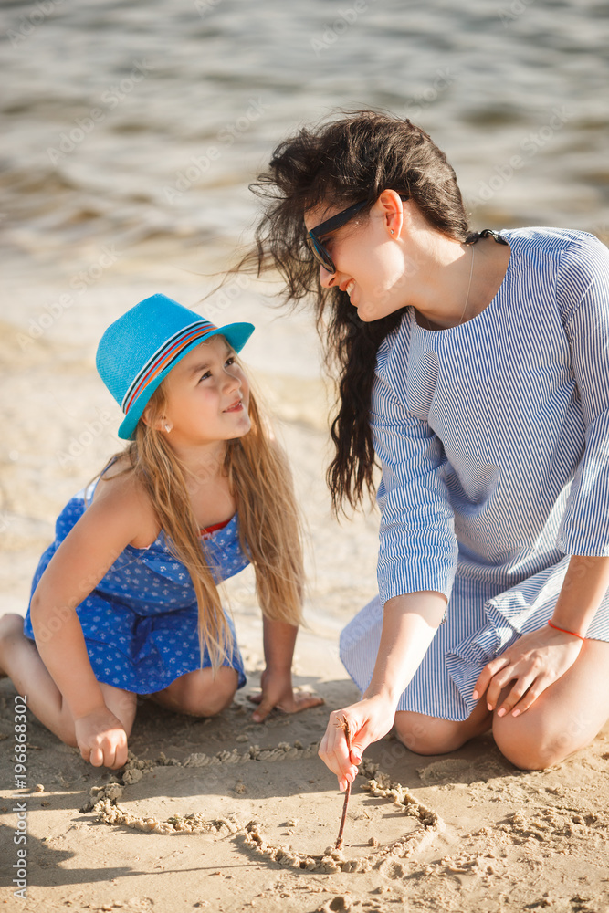Mother and her little daughter having fun at the coast. Young pretty mom and her child playing near the water and drawing heart on the sand Mother and her little daughter having fun at the coast. Young pretty mom and her child playing near the water and drawing heart on the sand