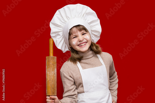 Portrait of happy little girl in chef uniform holds a rolling pin isolated on red. Kid chef. Cooking Process Concept 