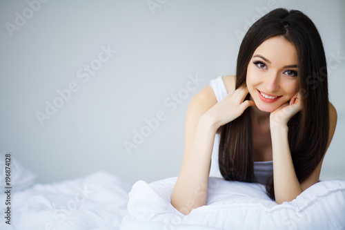 Care Concept. A beautiful young smiling brunette woman lying in white bed comfortably and blissfully.