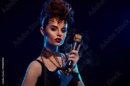 Young model with bright make up, including liner, long lashes. Female warrior wearing stylish hairdress with curles. Girl is wearing black top with thin chains. Woman holding steel sword with skull.