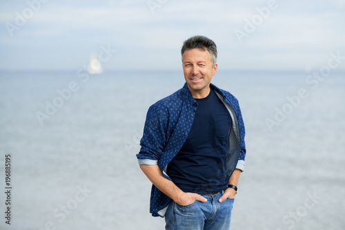 Handsome middle-aged man walking at the beach. Attractive mid adult male model posing at seaside in blue jeans, t-shirt shirt. Outdoor portrait of beautiful macho man.