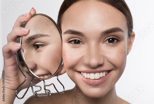 Natural attractiveness. Close up portrait if positive delighted young woman is standing and holding mirror near her face while reflected in it. She is looking at camera with joy. Isolated background