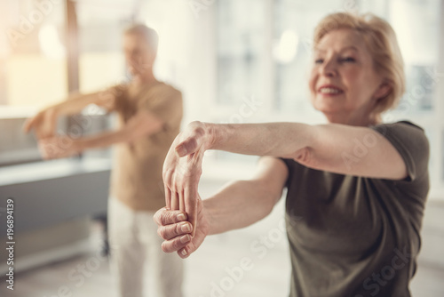 Joyful aging woman standing and stretching her hand. Old male in same posture standing on background. Focus on hands