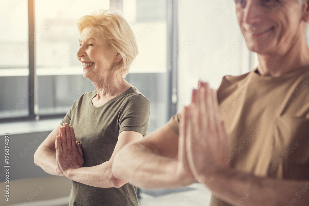 Delighted senior woman standing with hands clasped at her chest and smiling. Old man in same posture standing beside her. Focus on female Delighted senior woman standing with hands clasped at her chest and smiling. Old man in same posture standing beside her. Focus on female