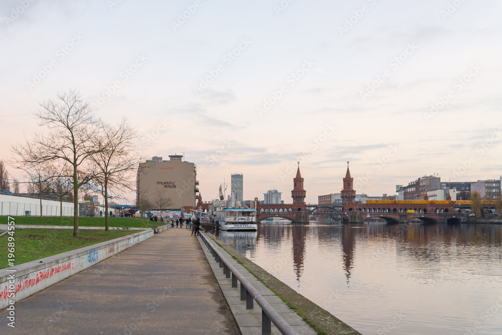 Scenery and pedestrian way of riverside along Spree river and East side gallery with Oberbaumbrücke behind during sunset in Berlin, Germany.