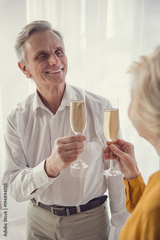Enjoyed retired man and woman standing in living room and going clinking glasses with champagne Enjoyed retired man and woman standing in living room and going clinking glasses with champagne