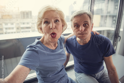 Portrait of joking elderly people playing the ape while making selfie near window indoors