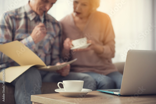 Focus on mug and laptop on table. Elderly couple focused on documents on background