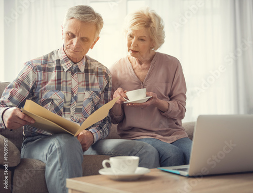 Calm mature man and woman sitting in living room and looking at folder with papers