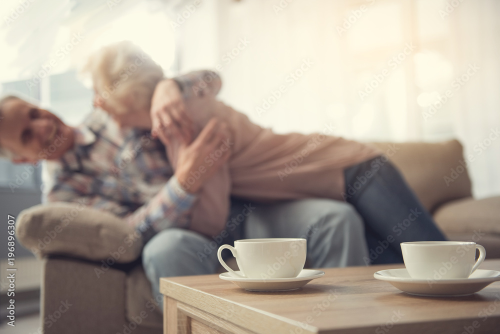 Focus on mugs on desk. Laughing old man and woman lying on sofa and hugging Focus on mugs on desk. Laughing old man and woman lying on sofa and hugging
