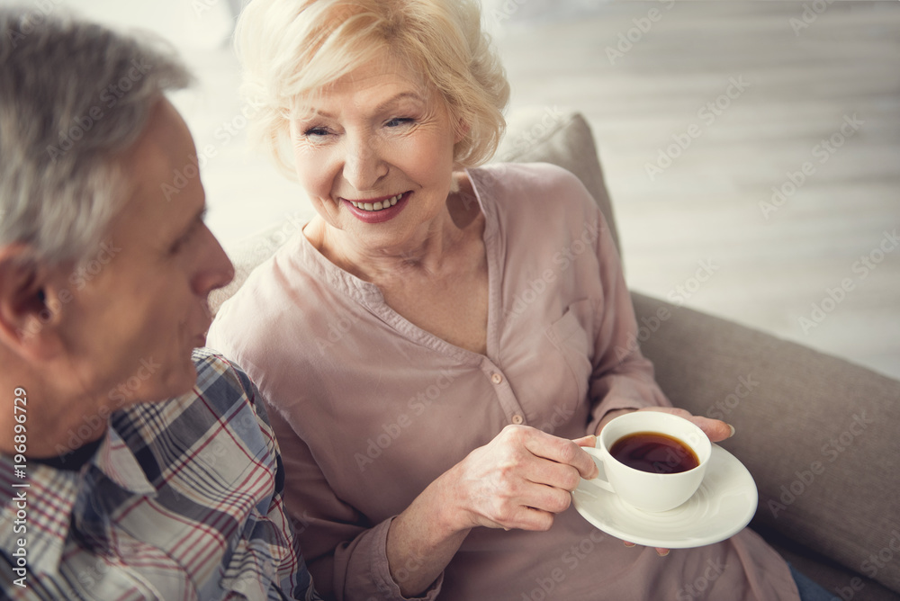 Smiling granny holding cup of tea while resting and talking with senior man indoors Smiling granny holding cup of tea while resting and talking with senior man indoors