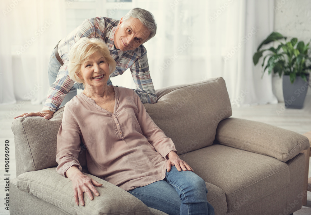 Portrait of glad pensioners spending time inside. Woman is sitting on sofa and man standing behind her back. They are looking at camera with joy. Copy space in right side