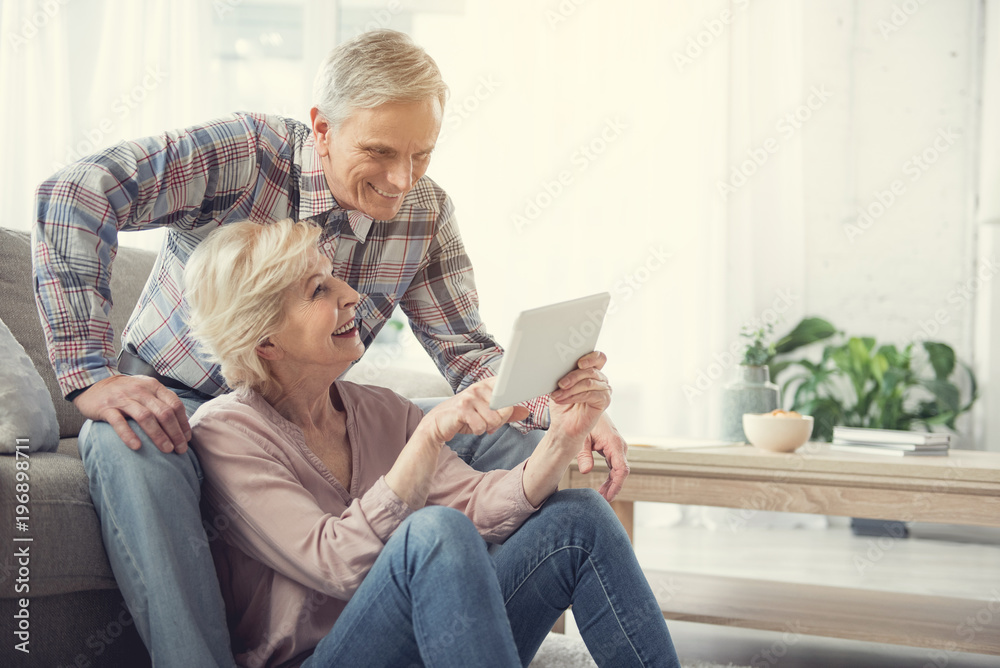 Happy old woman sitting on the floor and pointing at tablet to her husband. Glad man is resting on couch. Copy space in right side - obrazy, fototapety, plakaty 