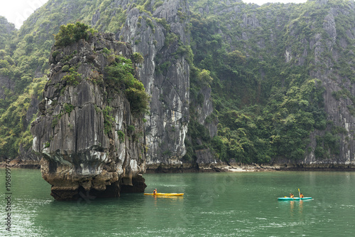 Enormous towering limestone islands over emerald water with tourists kayaking under the void of island in summer at Quang Ninh, Vietnam.