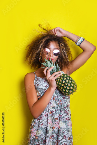 Fashion summer portrait smiling afro american girl in sunglasses and pineapple over yellow background