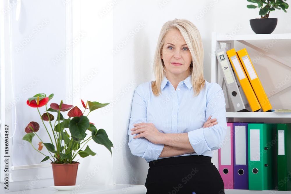 Successful business woman looking confident and smiling. business woman in blue shirt looking friendly into camera standing near window in office Successful business woman looking confident and smiling. business woman in blue shirt looking friendly into camera standing near window in office