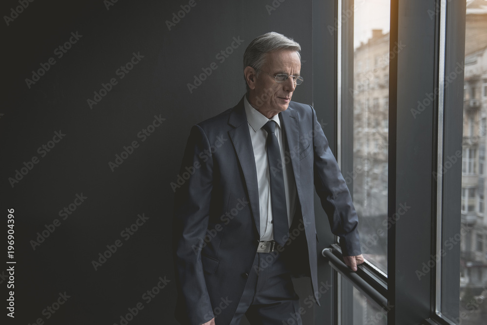 Portrait of pensive senior male looking at window while situating in room. Relax and labor concept
