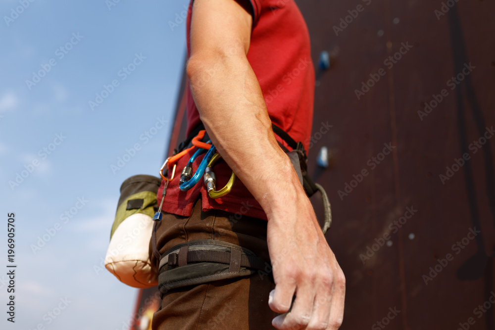 Close-up detail of rock climber wearing safety harness and climbing equipment outdoor wich chalk magnesium bag and carabiners. Artificial training climbing wall on background. Man prepares for workout Close-up detail of rock climber wearing safety harness and climbing equipment outdoor wich chalk magnesium bag and carabiners. Artificial training climbing wall on background. Man prepares for workout