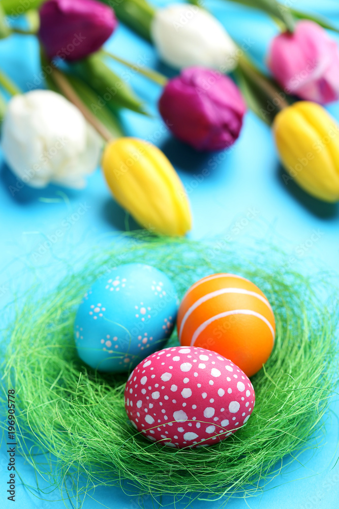 Colorful easter eggs in green nest with tulips on blue background Colorful easter eggs in green nest with tulips on blue background