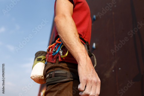 Close-up detail of rock climber wearing safety harness and climbing equipment outdoor wich chalk magnesium bag and carabiners. Artificial training climbing wall on background. Man prepares for workout