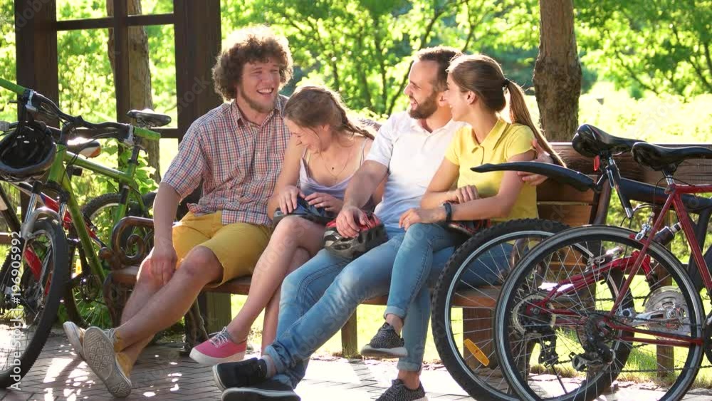 Young cheerful friends resting in park. Four cute cyclist sitting on bench in park, laughing and talking. Happy summer weekend. Young cheerful friends resting in park. Four cute cyclist sitting on bench in park, laughing and talking. Happy summer weekend.