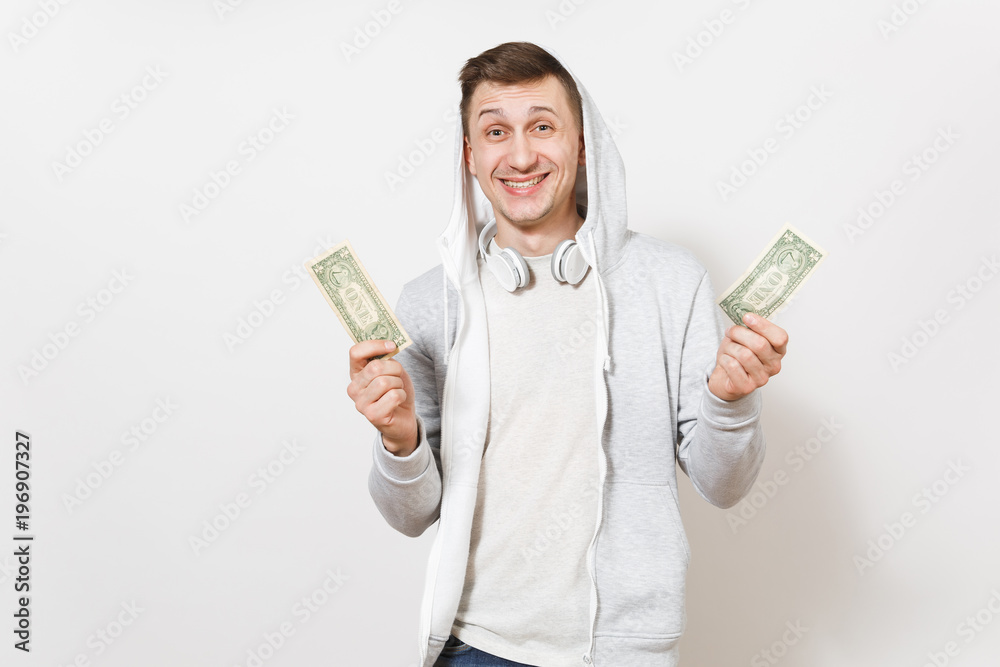 Young handsome smiling student in t-shirt, blue jeans and light sweatshirt with hood with headphones holds two one-dollar bills and rejoiced in studio on white background. Concept of emotions Young handsome smiling student in t-shirt, blue jeans and light sweatshirt with hood with headphones holds two one-dollar bills and rejoiced in studio on white background. Concept of emotions