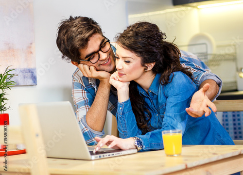 Young couple sitting by table in their apartment and looking at laptop.