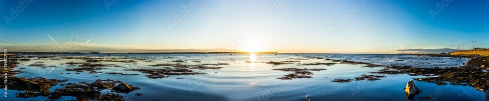Panorama of Rimouski sunset in Gaspesie region of Quebec, Canada with sun path, seaweed, rocks and shallow water, reflection